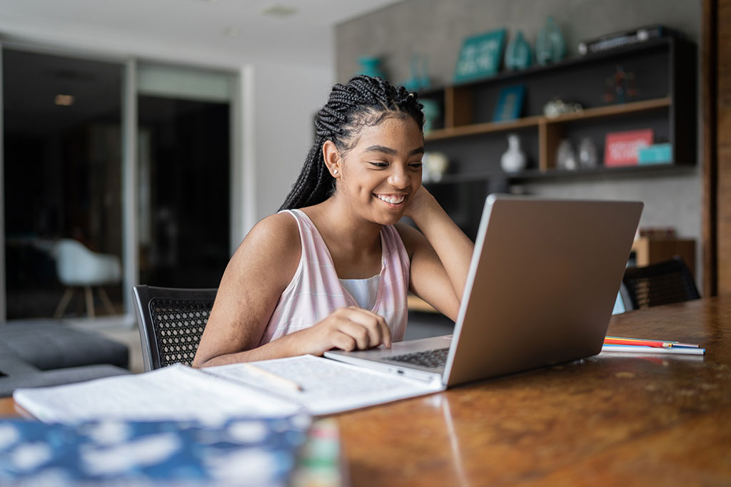 Student smiling while using a laptop at a table in a home setting, with notebooks and study materials nearby.